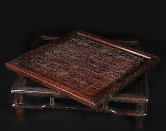 May include: A dark brown wooden game board featuring a grid pattern with etched lines, resting at an angle on a matching four-legged wooden stand. The surface of the square board displays intricate, thin, light-colored geometric markings.