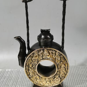 May include: A black and gold teapot with a bamboo handle. The teapot has a round body with a hole in the center and is decorated with raised figures around the edge.