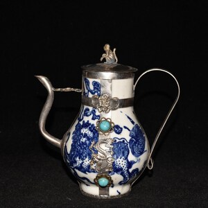 Könnte beinhalten: Eine weiße Porzellan-Teekanne mit einem blauen und weißen Drachenmuster. Die Teekanne hat einen silbernen Deckel und Griff und ist mit türkisfarbenen Steinen verziert.