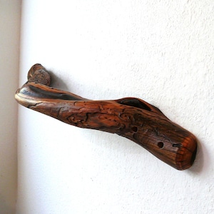 May include: A wall-mounted handle crafted from a piece of natural driftwood, featuring a rich, dark brown polished finish that highlights the organic curves, knots, and textured grain patterns of the wood, securely attached to a white wall.
