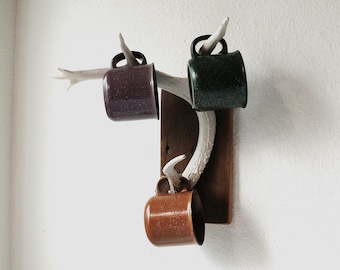 May include: A rustic wall-mounted mug rack featuring three speckled mugs in brown, green, and purple. The mugs are suspended from a deer antler attached to a wooden plaque. The rack is mounted on a white wall.