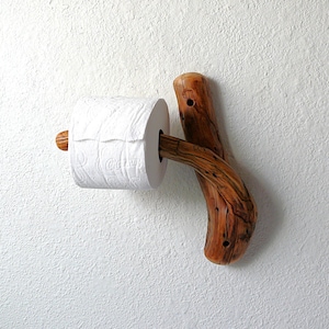 May include: A wall-mounted toilet paper holder crafted from a natural, curved piece of polished brown wood with visible grain and knots, holding a single roll of white toilet paper on its horizontal branch extension.