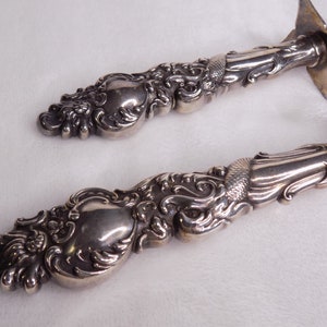 May include: A pair of ornate silver serving utensils with intricate scrollwork and a polished finish.
