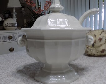 May include: A white ceramic soup tureen with a lid and a ladle. The tureen has a pedestal base and two ornate handles. The lid is topped with a decorative finial. The tureen is on a patterned tablecloth.