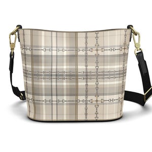 May include: A bucket bag featuring a beige and grey plaid pattern overlaid with a repeating equestrian bit and chain print. The bag includes a black adjustable shoulder strap attached with gold-toned metal hardware clips on both sides.