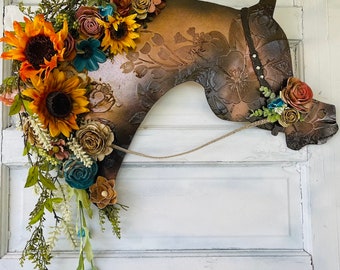 Può includere: Una silhouette di testa di cavallo marrone con un design floreale e un morso in pelle. La testa del cavallo è decorata con una varietà di fiori dai colori autunnali, tra cui girasoli, rose e bacche.