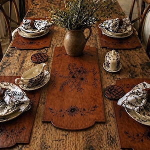 May include: A rustic dining table set for six. The table is adorned with brown leather placemats and a matching table runner, all featuring floral designs. Each place setting includes patterned plates, napkins, and a wooden napkin ring. A ceramic pitcher holds greenery.