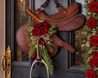May include: A decorative equestrian-themed door wreath featuring a brown leather saddle, adorned with red roses and greenery. A bronze horse head serves as the hanger. A matching rose garland frames the door. The door has a gold handle.