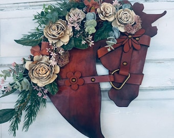 Puede incluir: Una corona de cabeza de caballo de cuero marrón adornada con un arreglo floral de flores crema y rosas, vegetación y pequeñas bayas marrones.