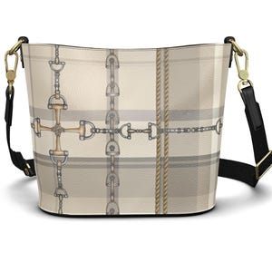 May include: A bucket bag featuring a beige and grey plaid pattern overlaid with a print of silver and gold-toned equestrian horse bits, stirrups, and rope details. The bag includes a black adjustable shoulder strap with gold-toned metal hardware.