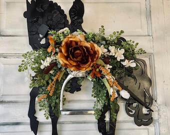 Può includere: Una ghirlanda a forma di cavallo in metallo nero con una silhouette di cavaliere, adornata con una grande rosa color ruggine, fiori bianchi e arancioni e fogliame verde. La ghirlanda include una staffa argentata ed è progettata per la decorazione di porte o pareti.
