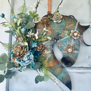 May include: A metal horse head silhouette with a teal, gold, and brown floral design. The horse head is decorated with rhinestones and is surrounded by a floral arrangement with blue, brown, and white flowers.