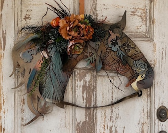 Può includere: Una decorazione da parete a forma di testa di cavallo. La testa di cavallo in metallo è adornata con fiori e piume nei toni del marrone, blu e arancione. Per appenderla viene utilizzato un cinturino in pelle. La testa di cavallo è montata su una porta bianca usurata.