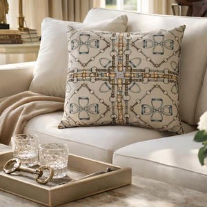 May include: A decorative square pillow with a beige background and a repeating equestrian bit pattern in shades of brown and blue. The pillow is placed on a white sofa, with a beige throw blanket. A gold tray with two glasses and a decorative bit is on the coffee table.