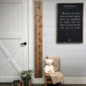 May include: A wooden growth chart with the words "The Zahn Family" and numbers 1 through 6. A black chalkboard sign with the words "The Sandlot" and a quote from the movie. A teddy bear sitting on a wooden chair with a blanket. Two potted plants with wooden stands.