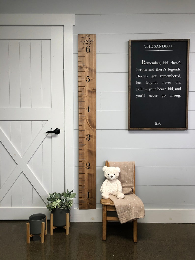 May include: A wooden growth chart with the words "The Zahn Family" and numbers 1 through 6. A black chalkboard sign with the words "The Sandlot" and a quote from the movie. A teddy bear sitting on a wooden chair with a blanket. Two potted plants with wooden stands.
