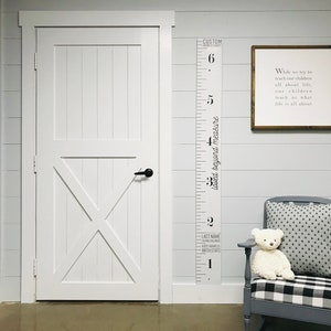 May include: A white wooden door with a black door handle, a white wooden growth chart with black numbers and text that reads "Loved beyond measure" and "Custom", a framed sign with the text "While we try to teach our children all about life, our children teach us what life is all about", a gray rocking chair with a black and white checkered cushion, and a white teddy bear sitting on the cushion.