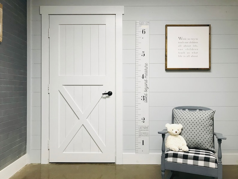 May include: A white wooden door with a black door handle, a white wooden growth chart with black numbers and text that reads "Loved beyond measure" and "Custom", a framed sign with the text "While we try to teach our children all about life, our children teach us what life is all about", a gray rocking chair with a black and white checkered cushion, and a white teddy bear sitting on the cushion.