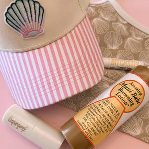 May include: A beige baseball cap with a pink and white striped brim and a seashell patch, a beige fabric with a seashell pattern, a white lip balm labeled BITCHSTIX, a Supergoop! stick, and an 8 fl. oz. bottle of Maui Babe Browning Lotion.