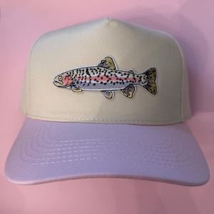 May include: A baseball cap featuring a cream-colored crown and a light purple brim. The front panel displays an embroidered patch of a rainbow trout with detailed scales, a pink lateral stripe, and yellow-tinted fins against a solid background.
