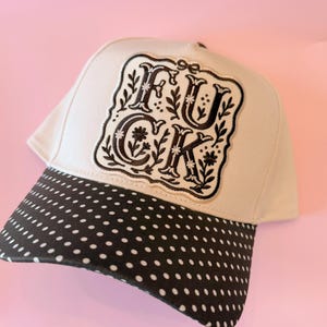 Puede incluir: Una gorra de béisbol color crema con un parche bordado en negro en el panel frontal. El parche muestra el texto FUCK en una fuente serif decorativa, rodeado de motivos florales y de enredaderas. La visera es negra con lunares blancos.