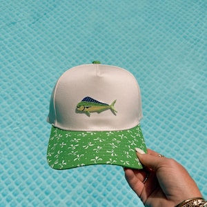 May include: A white baseball cap featuring an embroidered mahi-mahi fish in shades of blue, green, and yellow on the front panel. The brim is green with a repeating white floral or bow-like pattern. The cap has a structured crown and a top button.