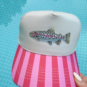 May include: A beige trucker hat featuring an embroidered rainbow trout patch on the front panel. The brim displays a vertical striped pattern in alternating shades of bright pink and light pink, with visible stitching along the edge of the brim.