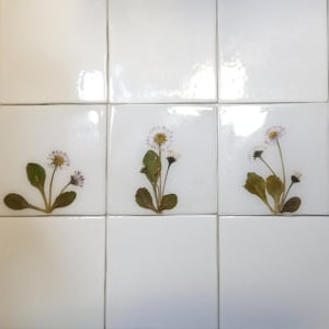 May include: A grid of nine square white ceramic tiles arranged in three rows and three columns. Three of the tiles feature pressed dried daisy flowers with green leaves and white petals, while the remaining six tiles are plain white.