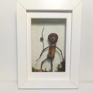 Op de afbeelding: Een ingelijst kunstwerk met een grillige figuur gemaakt van eikels en takjes. De figuur staat op een bed van mos en glitter en houdt een takje vast. De lijst is wit en heeft een glazen voorzijde.