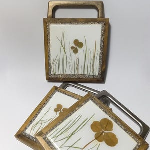 May include: Square coaster with a four-leaf clover and blades of grass encased in resin. The coaster has a wooden frame and a metal handle. The design is set against a white background.