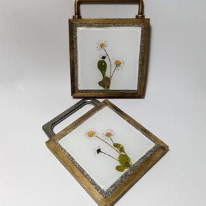 Puede incluir: Dos posavasos cuadrados con diseño floral. Cada posavasos presenta un fondo blanco con margaritas prensadas, hojas verdes y un marco dorado. Un posavasos tiene un asa. Los posavasos están hechos de madera y resina.