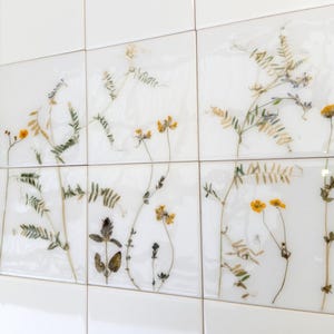 Puede incluir: Una pared de azulejos blancos con flores y hojas prensadas incrustadas en los azulejos. Las flores son amarillas y moradas.