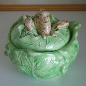 May include: A green ceramic cabbage shaped dish with a lid. The lid features three brown ceramic rabbits.