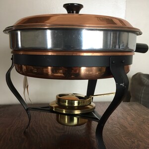 May include: A copper chafing dish with a black stand and a brass burner. The chafing dish has a silver lining and a black knob on the lid.