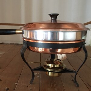 May include: Copper chafing dish with a black handle and a black metal stand with three legs. The dish has a lid and a burner underneath.