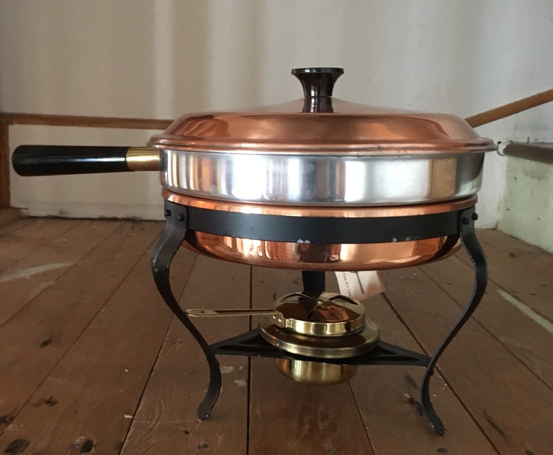 May include: Copper chafing dish with a black handle and a black metal stand with three legs. The dish has a lid and a burner underneath.