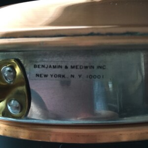 May include: Close-up of a copper pot with a black handle. The pot has a silver interior and a label that reads "Benjamin & Medwin Inc. New York, N.Y. 10001".