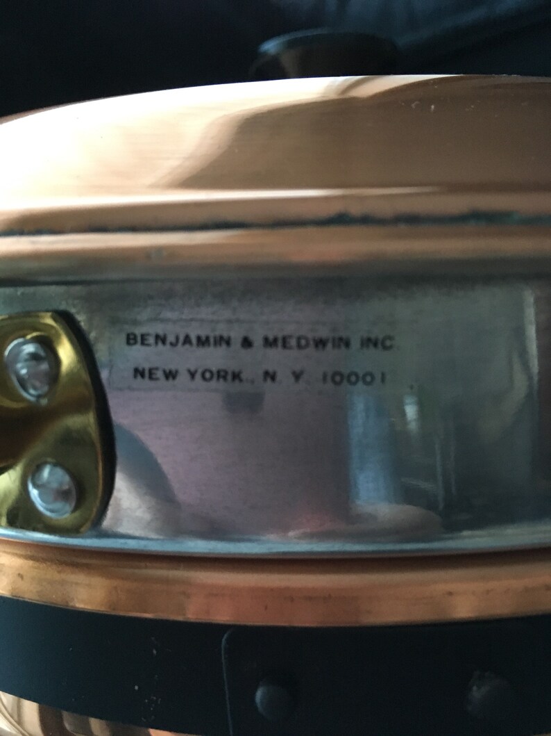 May include: Close-up of a copper pot with a black handle. The pot has a silver interior and a label that reads "Benjamin & Medwin Inc. New York, N.Y. 10001".