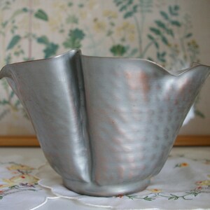 May include: A silver metal bowl with a wavy, ruffled edge. The bowl has a hammered finish and a slightly copper-toned hue.