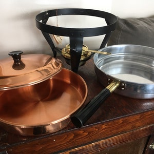 May include: A copper pot with a lid, a silver pot with a black handle, and a black metal stand with a gold burner.