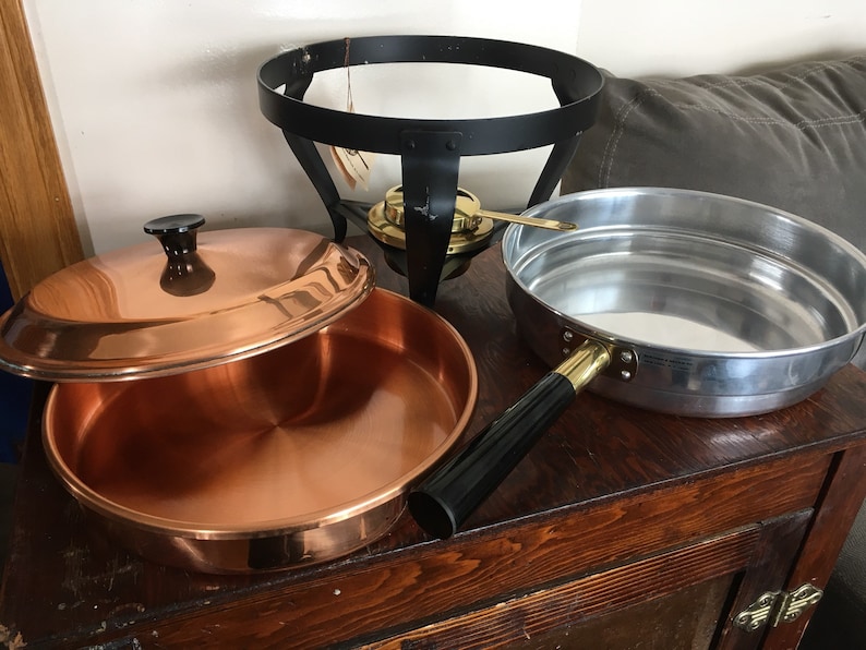 May include: A copper pot with a lid, a silver pot with a black handle, and a black metal stand with a gold burner.