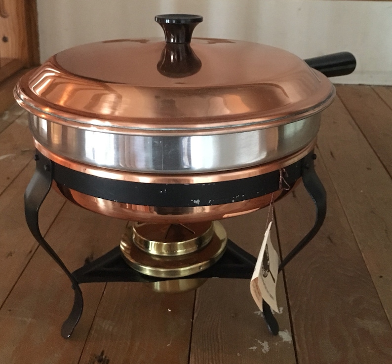 May include: Copper chafing dish with a black handle and a brass base. The chafing dish is on a wooden surface.