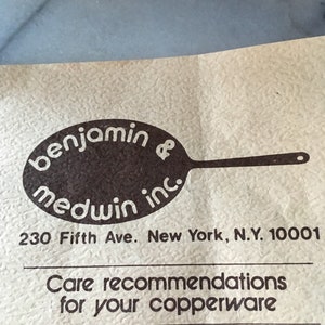 May include: A brown and white logo for Benjamin Medwin Inc. with the text "benjamin medwin inc." and a stylized image of a frying pan. The logo is on a beige paper with the address "230 Fifth Ave. New York, N.Y. 10001" and the text "Care recommendations for your copperware".