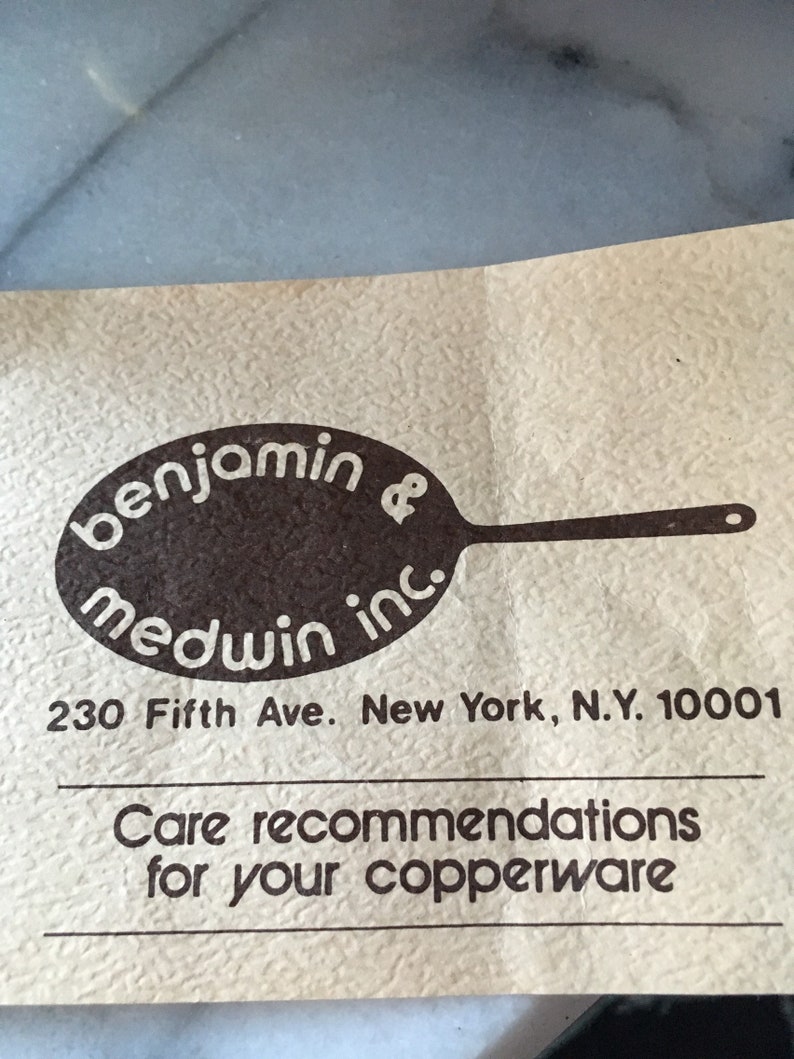 May include: A brown and white logo for Benjamin Medwin Inc. with the text "benjamin medwin inc." and a stylized image of a frying pan. The logo is on a beige paper with the address "230 Fifth Ave. New York, N.Y. 10001" and the text "Care recommendations for your copperware".