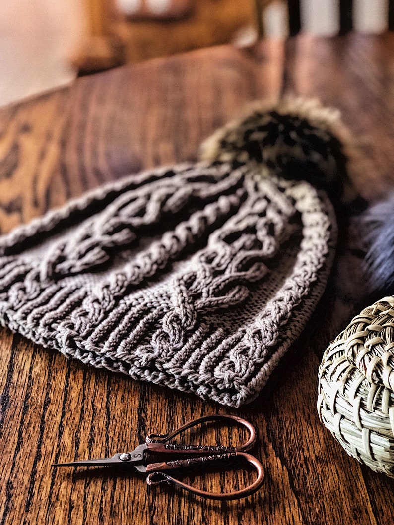 May include: A gray knitted hat with a pom pom on top, a pair of small scissors with a copper finish, and a woven basket on a wooden surface.
