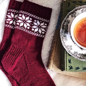 May include: A pair of red knitted socks with a white snowflake pattern on the cuff. The socks are lying on a white fuzzy surface next to a stack of books and a cup of tea.