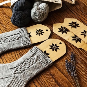 May include: Two gray knitted socks with a cable knit pattern, two balls of yarn in a fabric bag, and three wooden sock blockers with snowflake designs.