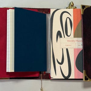 Puede incluir: Diario de cuero abierto con una cubierta roja, una inserci&oacute;n azul marino y un planificador semanal. El planificador tiene un dise&ntilde;o abstracto colorido y las palabras "Weekly Planner". Se ven esquinas doradas y una cadena.