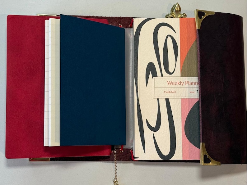 Puede incluir: Diario de cuero abierto con una cubierta roja, una inserci&oacute;n azul marino y un planificador semanal. El planificador tiene un dise&ntilde;o abstracto colorido y las palabras "Weekly Planner". Se ven esquinas doradas y una cadena.
