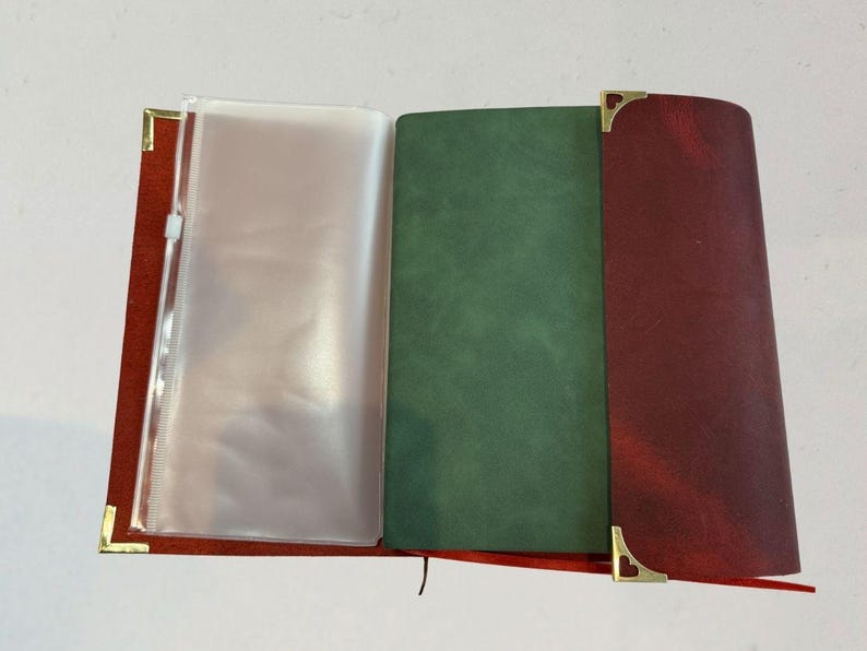 Puede incluir: Diario de cuero abierto con una cubierta roja, p&aacute;ginas interiores verdes y burdeos y una funda de pl&aacute;stico transparente. Las esquinas doradas a&ntilde;aden un toque decorativo. El diario est&aacute; abierto, mostrando las p&aacute;ginas interiores.
